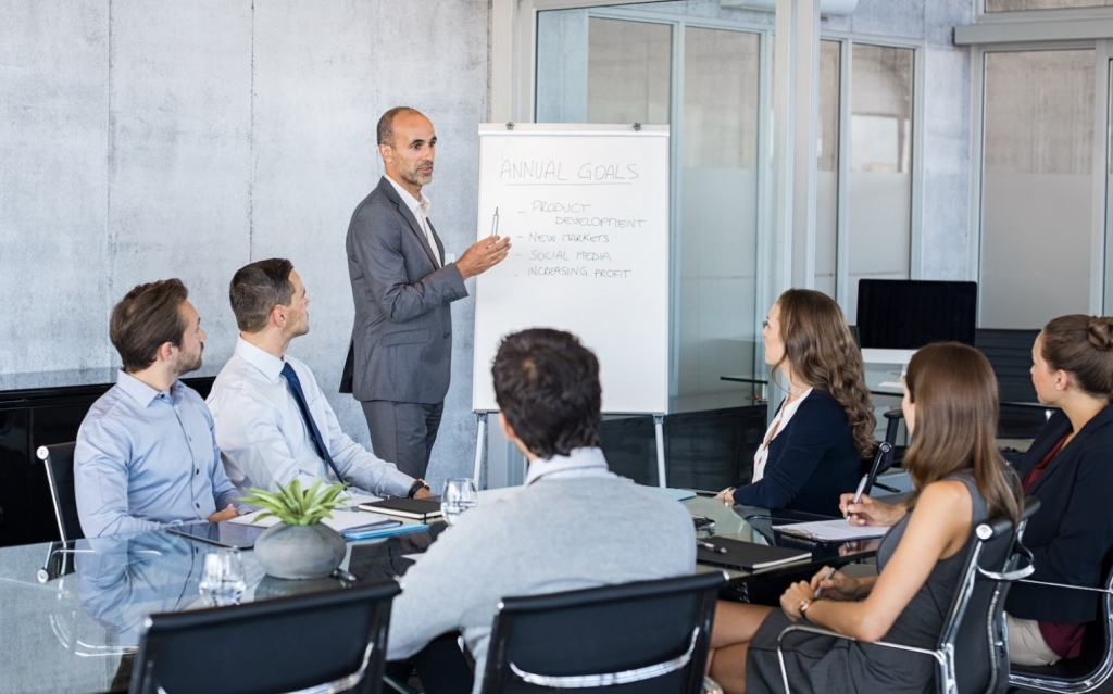 Confident mature businessman giving a presentation to his team in modern office. Business brief with annual goals with employees. Leadership man training businessmen and businesswomen in conference room.